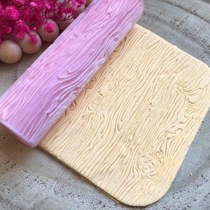 May include: A pink silicone rolling pin with a wood grain pattern is rolling over a beige fondant sheet with the same wood grain pattern.