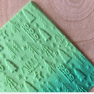 Può includere: Tappetino da forno in silicone verde a tema natalizio. Il tappetino presenta un motivo ripetitivo di alberi di Natale, stelle, fiocchi di neve e le parole "Ho Ho" e "Buon Natale".