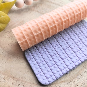 May include: A close-up of two silicone rolling pins with a brick pattern. The top rolling pin is peach colored and the bottom rolling pin is light purple.