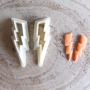 Può includere: Due tagliabiscotti a forma di fulmine in plastica bianca e due fulmini in pasta di zucchero arancione su una superficie di legno marrone chiaro.