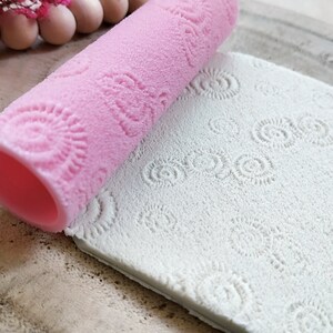 May include: A pink silicone rolling pin with a repeating pattern of small circles. The rolling pin is rolling over a white silicone mat with the same pattern embossed on it.