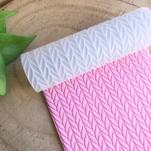 Könnte beinhalten: Eine weiße Silikon-Nudelrolle mit einem erhabenen Strickmuster, neben einer rosa Silikonmatte mit dem gleichen Muster.