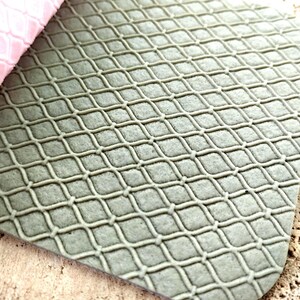 Puede incluir: Primer plano de una alfombra gris texturizada con un patrón en forma de diamante. Una sección de material rosa es visible en la esquina superior izquierda. La alfombra tiene un diseño similar a una cuadrícula, creando una superficie texturizada.