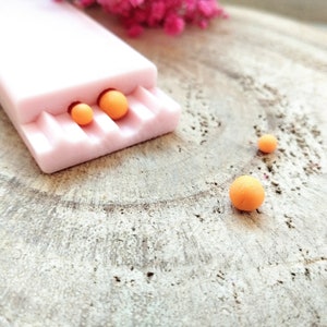 Könnte beinhalten: Eine rosa Plastikform mit drei runden orangefarbenen Perlen, zwei in der Form und eine auf einer Holzoberfläche.