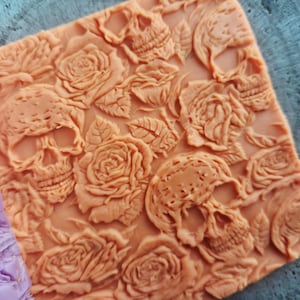 May include: A square, peach-colored soap bar with a carved design of skulls and roses. The skulls are interspersed with detailed rose blooms and leaves, creating a gothic floral pattern. The soap has a textured surface.