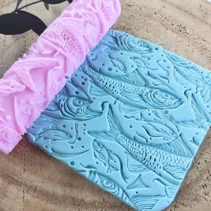 Puede incluir: Un rodillo rosa con un diseño de pescado y una estera de silicona azul con un diseño de pescado. El rodillo se utiliza para crear impresiones en fondant o masa.