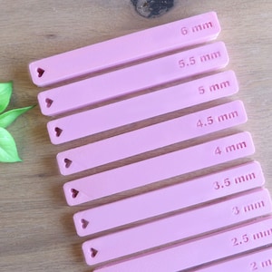 Könnte beinhalten: Neun rosa Plastikstreifen mit herzförmigen Ausschnitten und Maßen in Millimetern, von 2 mm bis 6 mm.