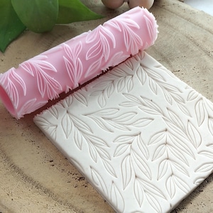 Pode incluir: Um rolo rosa com um padrão de folhas ao lado de uma telha de argila branca com um desenho de folhas em relevo. O rolo é cilíndrico e apresenta um padrão de folhas repetido. A telha tem um padrão de folhas semelhante.