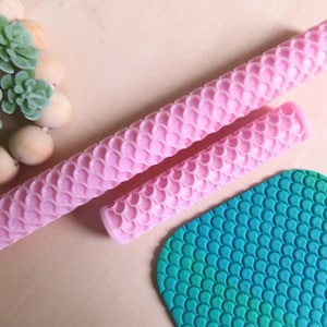 Puede incluir: Dos rodillos de amasar de silicona rosa con un patrón de escamas. Los rodillos de amasar están sobre una superficie marrón claro. Una lámina de fondant verde y azul con un patrón de escamas se encuentra en la esquina inferior derecha de la imagen.