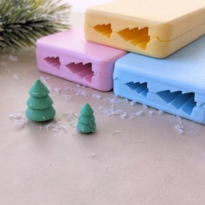 May include: Three pastel-colored plastic molds for making Christmas tree-shaped decorations. The molds are stacked on top of each other, with the top mold being yellow, the middle mold being pink, and the bottom mold being blue. Two small green Christmas trees made with the molds are in the foreground.