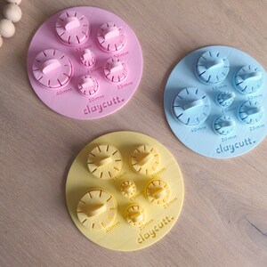 Può includere: Tre set di tagliapasta rotondi in plastica, rosa, blu e giallo. Ogni set ha sei tagliapasta di diverse dimensioni, che vanno da 15 mm a 40 mm. I tagliapasta sono etichettati con la loro dimensione in millimetri. I set sono disposti in cerchio su una superficie di legno. Il testo "claycutt." è stampato su ogni set.