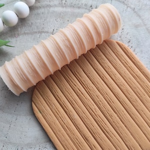 May include: A beige silicone rolling pin with a ribbed texture, used for creating wood grain patterns on fondant or clay. The rolling pin is lying on a brown textured surface.