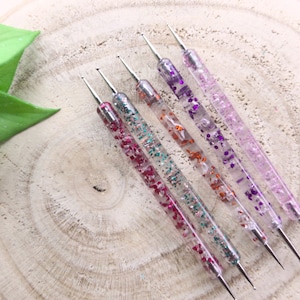 以下が含まれることがあります： ハンドルにグリッターが埋め込まれた透明なネイルアートドットツール5本セット。ツールは、パープル、オレンジ、グリーンなど、さまざまな色のグリッターを使用しています。