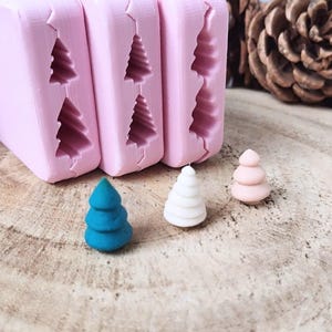 Könnte beinhalten: Rosa Silikonformen mit Weihnachtsbaumformen, neben drei Miniatur-Baumfiguren in Blau, Weiß und Rosa. Die Formen sind rechteckig, und die Bäume stehen auf einer Holzoberfläche.