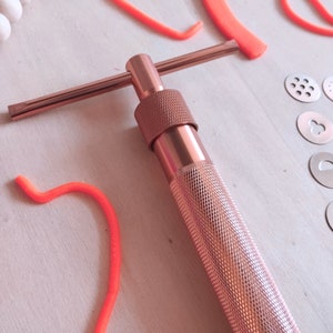 Peut inclure: Un outil en m&eacute;tal rose gold avec une poign&eacute;e en forme de T et un long baril cylindrique. L'outil est utilis&eacute; pour faire des formes d&eacute;coratives dans le fondant ou l'argile.