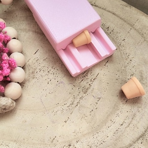 Peut inclure: Un récipient rectangulaire rose clair avec un couvercle, contenant deux petits pots de fleurs miniatures de couleur beige. Le récipient a des rainures à l'intérieur. Des fleurs roses séchées et des perles en bois sont en arrière-plan.