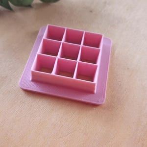 May include: A pink square cookie cutter with nine equally sized square compartments. The cutter has a raised edge with a textured pattern. The tool is designed for baking and crafting, perfect for creating uniform shapes.