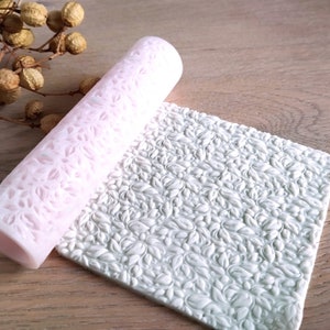 Pode incluir: Um rolo de silicone rosa com um padrão de folhas, e um tapete de silicone branco com um padrão de folhas em relevo.