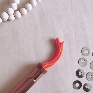Peut inclure: Gros plan sur un outil en cuivre pour la cr&eacute;ation de d&eacute;corations en argile. L'outil poss&egrave;de un manche et une buse remplie d'argile orange. La buse est fix&eacute;e &agrave; un tube m&eacute;tallique qui est reli&eacute; au manche. L'outil est sur une surface blanche.