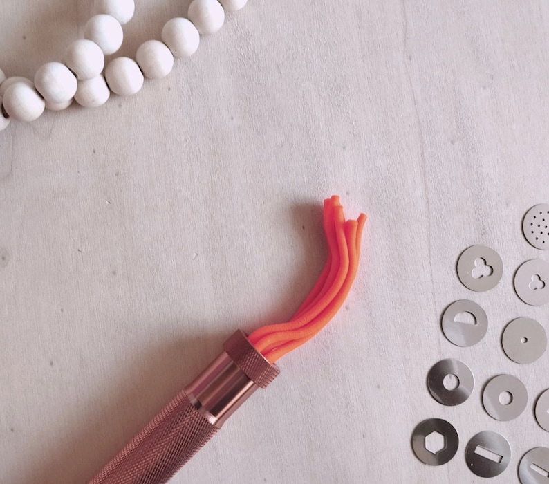 Peut inclure: Gros plan sur un outil en cuivre pour la cr&eacute;ation de d&eacute;corations en argile. L'outil poss&egrave;de un manche et une buse remplie d'argile orange. La buse est fix&eacute;e &agrave; un tube m&eacute;tallique qui est reli&eacute; au manche. L'outil est sur une surface blanche.