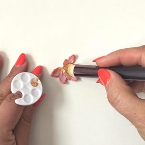 Puede incluir: Una mano sostiene una pequeña paleta blanca con pintura dorada en uno de los pozos. Otra mano sostiene un pincel y está aplicando la pintura dorada a una flor de arcilla rosa y naranja.