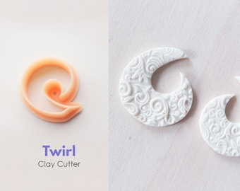 Peut inclure: Un emporte-pièce en plastique orange clair en forme de spirale, avec les mots "Twirl Clay Cutter" imprimés en dessous. Deux pièces d'argile blanches en forme de croissant de lune avec un motif floral détaillé sont visibles sur le côté droit de l'image.