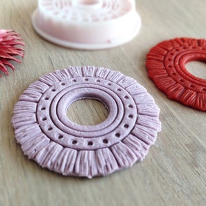 Könnte beinhalten: Zwei runde, dekorative Ornamente aus Polymer Clay. Eines ist helllila mit einem strukturierten, gefranstem Rand und einem Kreisdesign in der Mitte. Das andere ist einfarbig rot mit einem ähnlichen gefranstem Rand und Kreisdesign.