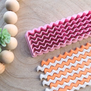 Könnte beinhalten: Ein rosa-farbener, wellenförmiger Fondant-Ausstecher aus Kunststoff mit einer Musterprobe aus weißer und orangefarbener Fondant.