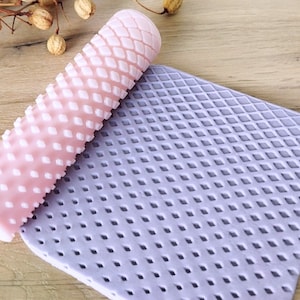Puede incluir: Un rodillo texturizado rosa y blanco con un patrón de diamante, y una estera de silicona púrpura con un patrón de diamante.