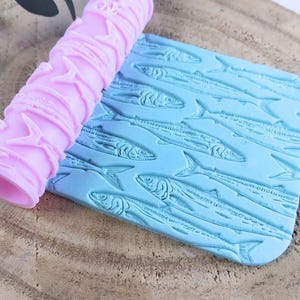 Puede incluir: Un rodillo de silicona rosa con un diseño de peces en relieve. El rodillo está sobre una estera de silicona azul con un diseño de peces en relieve.