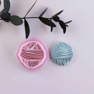 Op de afbeelding: Een roze bolvormige mal naast een lichtblauwe bol. De roze mal heeft een rode bolvormige indruk. De bolvormen staan op een wit oppervlak met een donkergroene, bladrijke tak op de achtergrond.