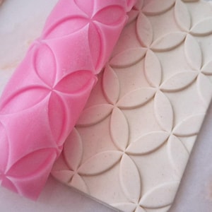 May include: A pink rolling pin with a floral pattern is shown next to a piece of off-white clay with the same embossed design. The rolling pin is a vibrant pink, and the clay has a natural, neutral tone. The pattern consists of overlapping circles.