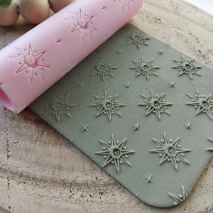 May include: A pink silicone rolling pin with a starburst pattern is rolled over a gray silicone mat with the same starburst pattern. The mat is textured with raised starbursts.