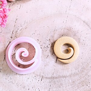 May include: Two cookie cutters in the shape of a spiral. One is pink and the other is beige.
