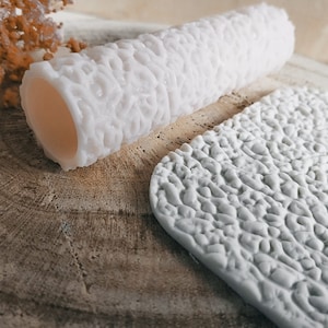 May include: A white silicone roller with a textured surface, used for creating a textured effect on clay or fondant. The roller is shown next to a flat piece of textured clay.