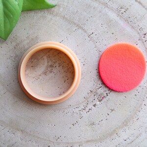 May include: A close-up of a round, light brown, plastic ring with a smooth surface. The ring is sitting on a light brown wooden surface. Next to the ring is a bright pink, round, felt circle.