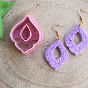 Puede incluir: Un par de pendientes morados con un diseño floral detallado. Los pendientes están hechos de arcilla polimérica y tienen ganchos dorados.