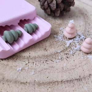 May include: A pink plastic mold with two green Christmas tree shaped objects inside. Two pink Christmas tree shaped objects are on a wooden surface with white flakes.