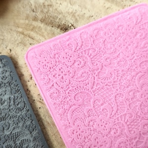 May include: Close-up of two silicone molds with intricate lace patterns. One mold is a soft pink, the other is gray. The molds are rectangular and feature detailed floral and geometric designs, perfect for crafting or baking.