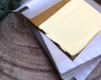 Può includere: Un mazzo di fogli di oro, ogni foglio è protetto da un sottile foglio di carta bianca. La foglia d'oro è di un colore oro brillante e lucido.
