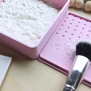 Puede incluir: Una estera de limpieza para brochas de maquillaje rosa con polvo blanco en un recipiente rosa. La estera tiene una cuadrícula de agujeros y el texto "cleanit" impreso en ella. Una brocha de maquillaje negra está sobre la estera.