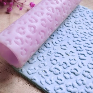 Pode incluir: Um rolo de silicone de cor lilás claro com um design de estampa de leopardo. O rolo está rolando sobre um tapete de silicone azul com o mesmo design de estampa de leopardo.