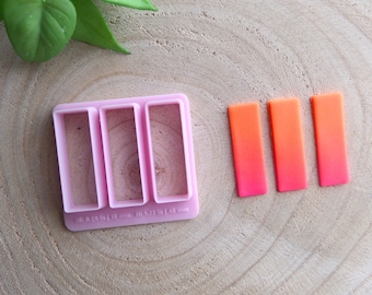 May include: Pink plastic mold with three rectangular cutouts for making polymer clay bars. The mold measures 1.5 x 2.7 inches. Three orange and pink gradient polymer clay bars are shown next to the mold.