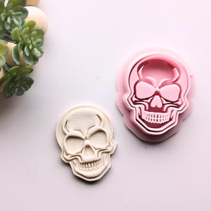 May include: Two skull-shaped cookie cutters, one pink and one white. The pink cutter is a raised design and the white cutter is a flat design.