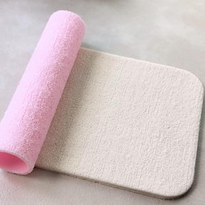 Könnte beinhalten: Eine aufgerollte rosa Badematte mit einer weißen Badematte darunter. Die Badematte besteht aus einem weichen, saugfähigen Material.