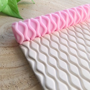 Puede incluir: Rodillo de amasar rosa con un patrón ondulado. El rodillo está sobre una estera de silicona blanca con un patrón ondulado.