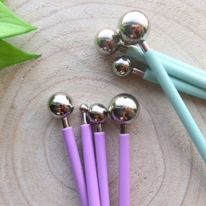 Puede incluir: Conjunto de herramientas con puntas de bola con mangos morados y azul claro. Las herramientas se utilizan para manualidades y la creación de patrones en arcilla o fondant.