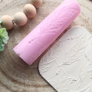Könnte beinhalten: Ein rosa Silikon-Nudelholz mit einem detaillierten Federmuster. Das Nudelholz liegt neben einer weißen Silikonmatte mit einem sich wiederholenden Federmuster.