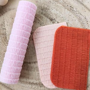 May include: Three pieces of textured paper in pink, light pink, and orange. The paper is in a ribbed pattern and is used for crafting.