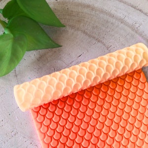 Puede incluir: Un rodillo de silicona con un patrón de escamas de sirena. El rodillo es naranja y blanco y está hecho de silicona. El rodillo está sobre una superficie de madera.